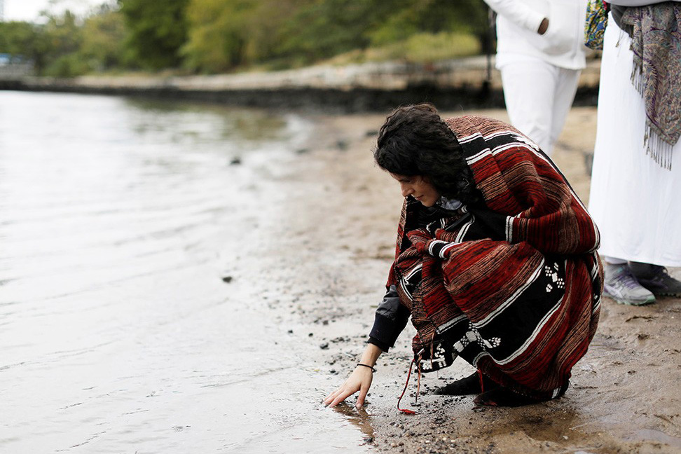 A woman by a lake bends down to touch the water. 